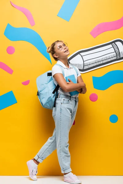 Full length view of dreamy schoolgirl looking up while holding book on yellow background with abstract elements and paper pencil — Fotografia de Stock