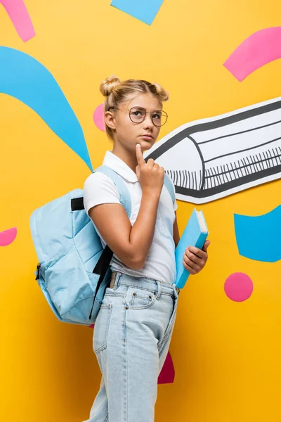 Thoughtful pupil with backpack holding book and looking away on yellow background with paper pencil and abstract elements — Stock Photo