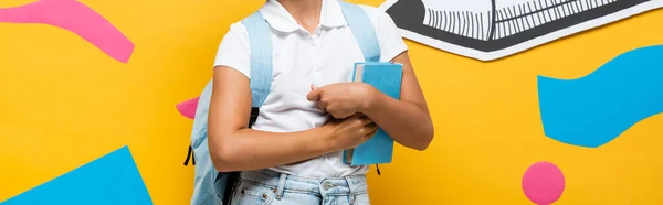 Vista recortada de la colegiala con la mochila sosteniendo libro cerca de lápiz de papel y elementos decorativos en amarillo, concepto panorámico - foto de stock