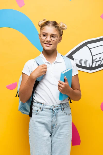 Skeptical schoolgirl in eyeglasses holding book on yellow background with paper pencil and multicolored elements — Stock Photo