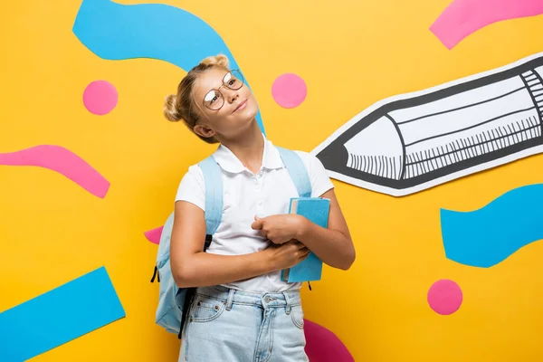 Verträumtes Schulmädchen mit Brille schaut weg, während sie ein Buch auf gelbem Hintergrund mit Papierstift und abstrakten Elementen in der Hand hält — Stockfoto