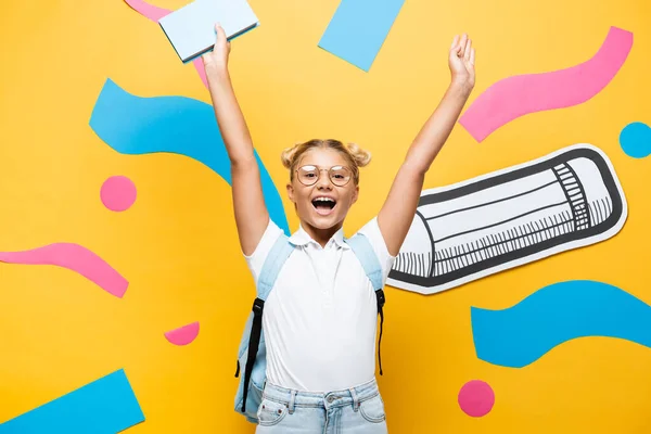 Excited schoolgirl in eyeglasses screaming while holding book in raised hands near paper pencil and decorative elements on yellow — Stock Photo
