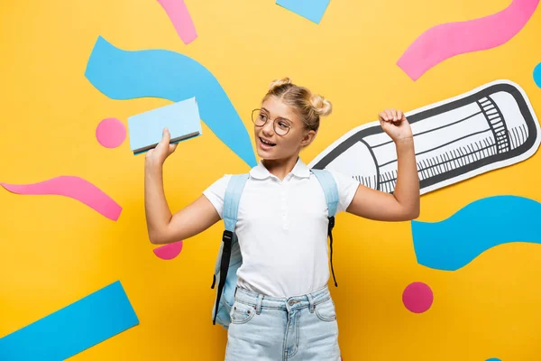 Взволнованная школьница в очках, держащая книгу и показывая жест триумфа возле бумажного карандаша и красочные элементы на желтом — стоковое фото