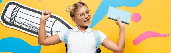 Panoramic shot of excited schoolgirl in eyeglasses holding book and showing winner gesture near colorful paper artwork on yellow — Stock Photo