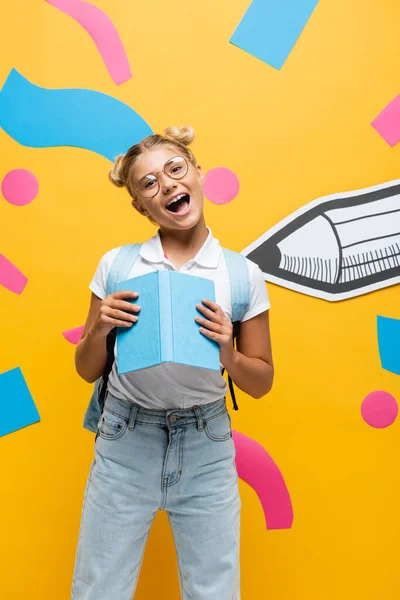 Excited schoolgirl in eyeglasses looking at camera with open mouth while holding book near paper pencil and decorative elements on yellow — Stock Photo