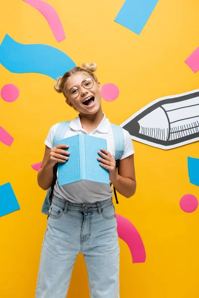 Aufgeregtes Schulmädchen mit Brille, das mit offenem Mund in die Kamera blickt, während es Buch in der Nähe von Papierstift und dekorativen Elementen auf gelb hält — Stockfoto