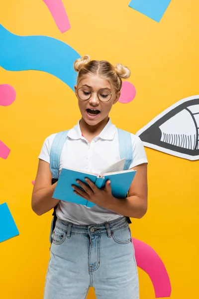 Excited schoolgirl in eyeglasses reading book on yellow background with paper pencil and multicolored elements — Stock Photo