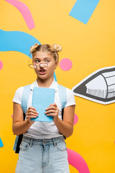 Bemused schoolgirl in skewed eyeglasses holding book near paper pencil and colorful elements on yellow — Stock Photo