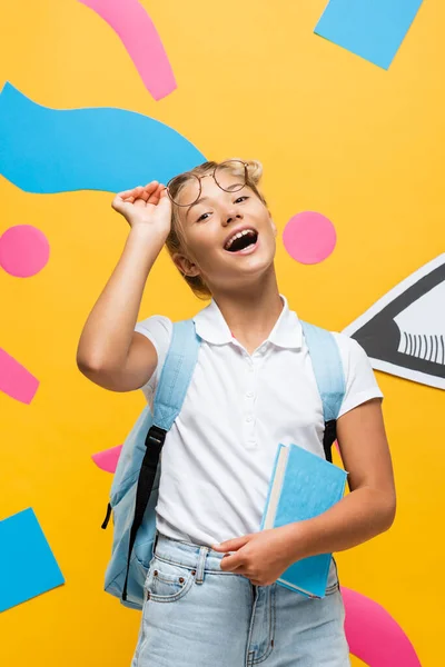 Aufgeregtes Schulmädchen berührt Brille, während sie Buch auf gelbem Hintergrund mit Papierstift und dekorativen Elementen hält — Stockfoto