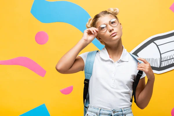 Scolaretta premurosa che tocca gli occhiali e distoglie lo sguardo vicino alla matita di carta tagliata ed elementi astratti sul giallo — Stock Photo