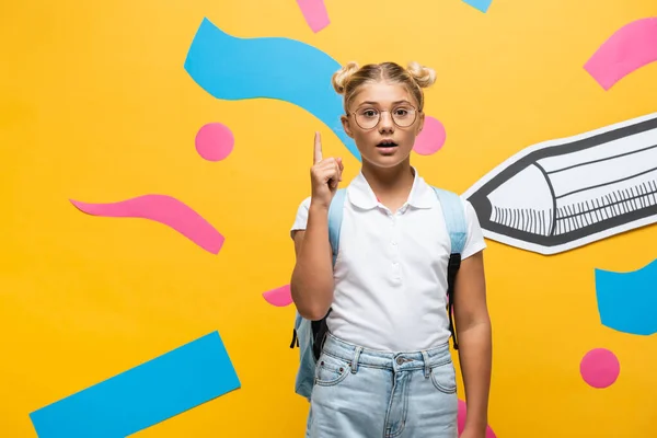 Schockierte Schülerin in Brille zeigt mit Finger auf dekorative Elemente und Bleistift auf gelb — Stockfoto