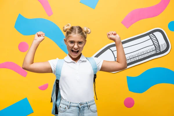 Excited schoolgirl in eyeglasses showing winner gesture near paper pencil and multicolored elements on yellow — Stock Photo