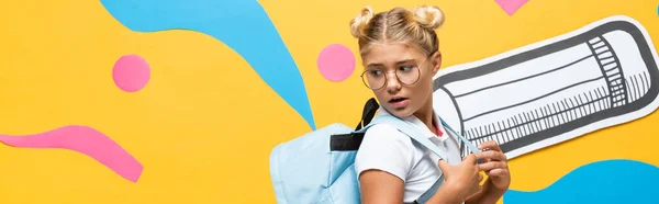 Imagen horizontal del colegial sorprendido mirando la mochila cerca de lápiz de papel y elementos decorativos en amarillo - foto de stock