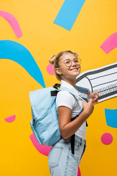 Радостная школьница с рюкзаком глядя в сторону бумажного карандаша и красочные элементы на желтый — стоковое фото