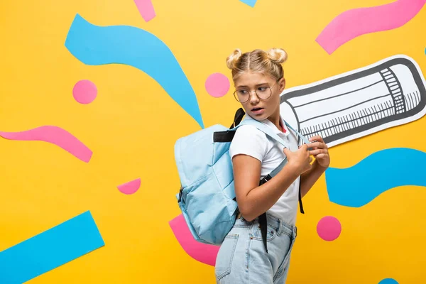 Alumno sorprendido mirando mochila sobre fondo amarillo con lápiz de papel y elementos abstractos - foto de stock