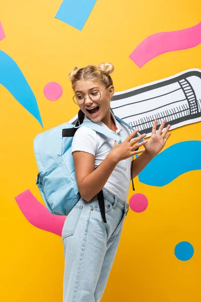 Erstauntes Schulkind blickt auf Rucksack neben Bleistift und bunten Elementen auf gelb — Stockfoto