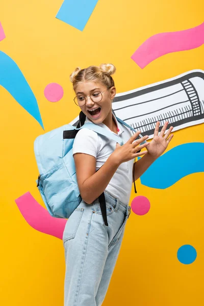 Scolaro stupito guardando lo zaino vicino alla matita di carta ed elementi multicolori sul giallo — Foto stock