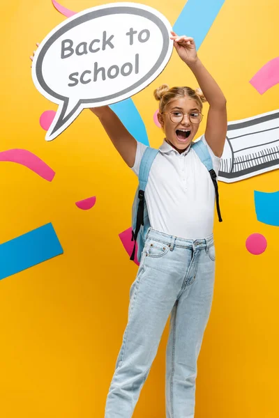 Aufgeregtes Schulmädchen schreit, während es eine Sprechblase mit dem Rücken zur Schule in der Nähe von Papierkunst und Bleistift auf gelb hält — Stockfoto