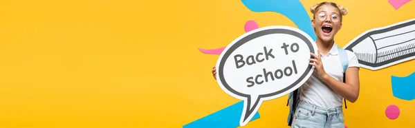 Panoramic concept of schoolgirl shouting while holding speech bubble with back to school inscription near colorful paper artwork on yellow — Stock Photo