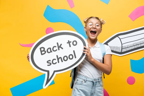 Aufgeregtes Schulmädchen schreit, während es eine Sprechblase mit dem Rücken zur Schule in der Nähe von Bleistift und bunten Elementen auf gelb hält — Stockfoto