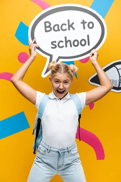 Emocionado colegial gritando mientras sostiene la burbuja del habla con la inscripción de vuelta a la escuela cerca de lápiz de papel y elementos abstractos en amarillo - foto de stock