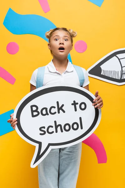 Sorprendido colegial mirando a la cámara mientras sostiene la burbuja del habla con letras de vuelta a la escuela sobre fondo amarillo con lápiz de papel y elementos coloridos - foto de stock