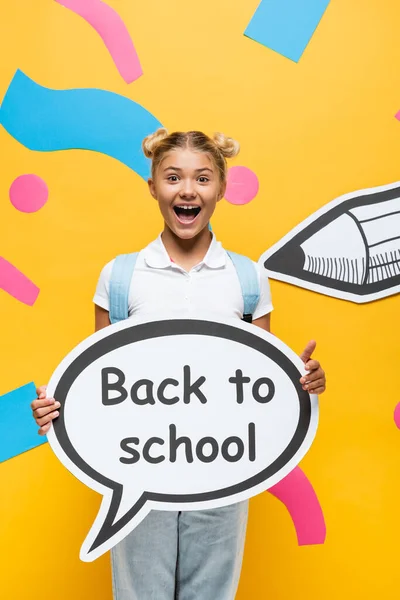 Excited schoolchild shouting while holding speech bubble with back to school inscription near abstract paper elements and pencil on yellow — Stock Photo