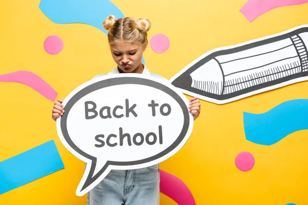 Displeased schoolgirl holding speech bubble with back to school inscription near paper pencil and decorative elements on yellow — Stock Photo