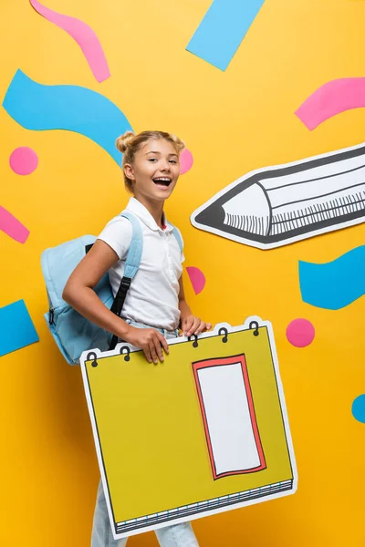 Criança da escola alegre olhando para a câmera enquanto segurando maqueta de livro de cópia perto de lápis de papel e elementos abstratos no amarelo — Fotografia de Stock