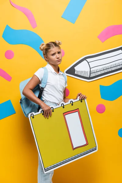 Displeased pupil looking at camera while holding notebook maquette on yellow background with paper pencil and decorative elements — Stock Photo