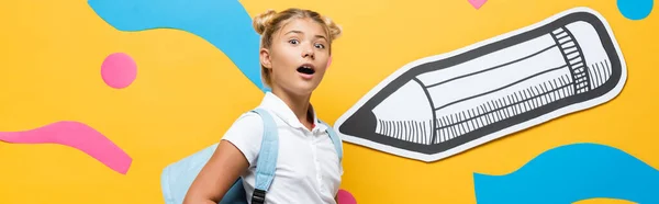 Horizontal concept of amazed pupil looking at camera on yellow background with paper pencil and multicolored elements — Stock Photo