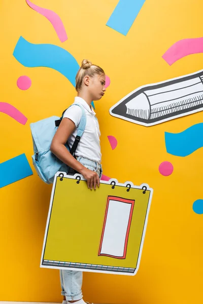 Side view of upset schoolgirl holding notebook maquette on yellow background with paper pencil and decorative elements — Stock Photo