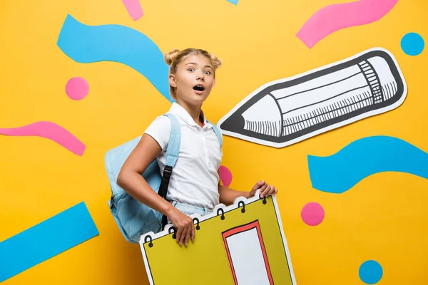 Surprised pupil with notebook maquette looking at camera near paper cut pencil and decorative elements on yellow — Stock Photo