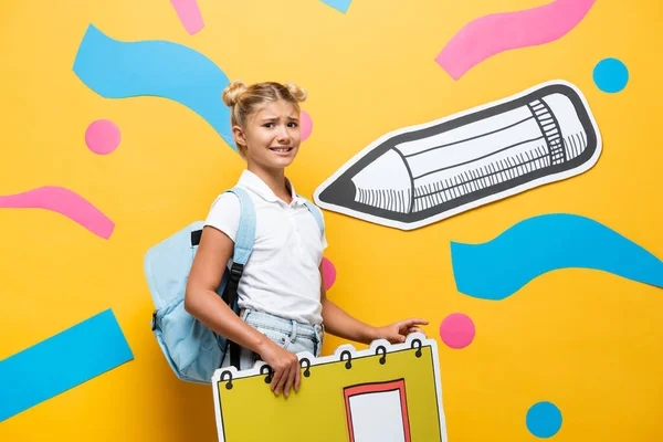 Angespannte Pupille mit Notizbuch-Maquette, die auf gelbem Hintergrund mit Bleistift und bunten Elementen in die Kamera blickt — Stockfoto