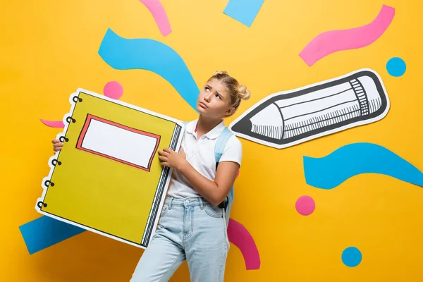 Nachdenkliche Pupille mit Kopierbuchmaquette auf gelbem Hintergrund mit dekorativen Elementen und Bleistift — Stockfoto