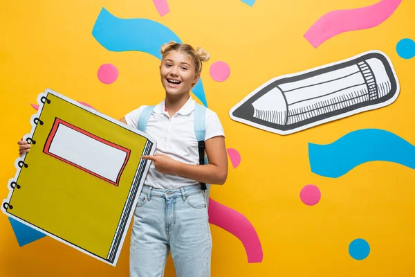 Excited pupil pointing with finger at notebook maquette on yellow background with paper cut pencil and abstract elements — Stock Photo