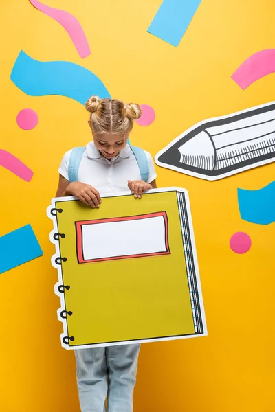 Aluna alegre segurando maquete de livro de cópia no fundo amarelo com lápis de corte de papel e elementos decorativos — Fotografia de Stock