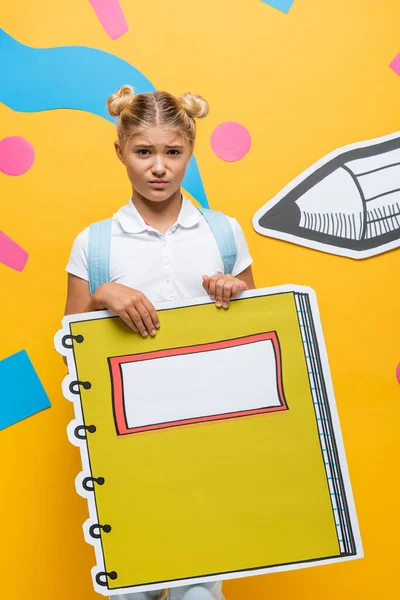 Aufgebrachte Schülerin mit Notizbuch-Maquette mit Blick auf Kamera auf gelbem Hintergrund mit Bleistift und abstrakten Elementen — Stockfoto