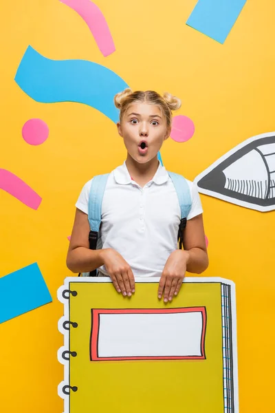 Schockiertes Schulmädchen mit Notizbuch-Maquette blickt in die Kamera auf gelbem Hintergrund mit Bleistift und bunten Elementen — Stockfoto