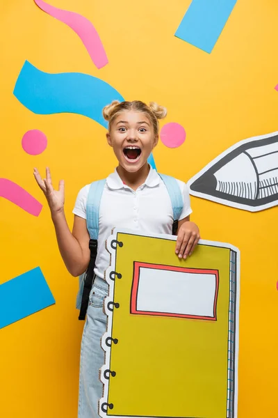 Excited schoolgirl with notebook maquette screaming and gesturing on yellow background with decorative elements and paper cut pencil — Stock Photo