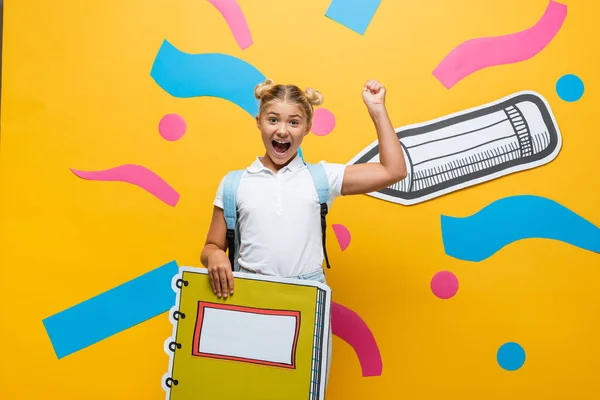 Alumno emocionado con maqueta libro copia mostrando gesto ganador y gritando sobre fondo amarillo con elementos decorativos y lápiz de corte de papel - foto de stock