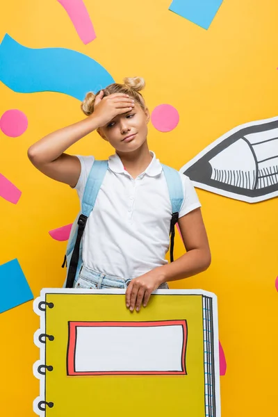 Exhausted pupil touching forehead while holding notebook maquette on yellow background with paper cut pencil and abstract elements — Stock Photo