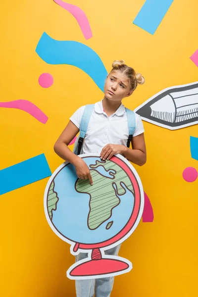 Upset schoolgirl holding globe maquette and looking away on yellow background with paper cut pencil and decorative elements — Stock Photo