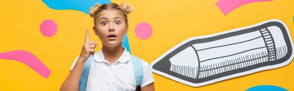 Horizontal crop of shocked pupil pointing with finger while looking at camera near paper pencil and decorative elements on yellow — Stock Photo
