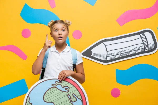 Erstaunt Schulkind mit Globus-Maquette zeigt Idee Geste und Blick in die Kamera in der Nähe von Papier Bleistift und abstrakte Elemente auf gelb — Stockfoto