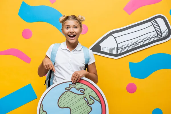 Joyeux écolier pointant du doigt la maquette globe près du crayon de papier et des éléments multicolores sur jaune — Photo de stock