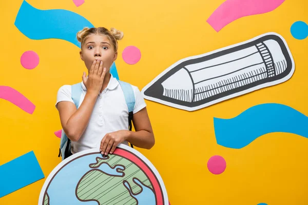 Shocked pupil covering mouth with hand while holding globe maquette near paper pencil and decorative elements on yellow — Stock Photo