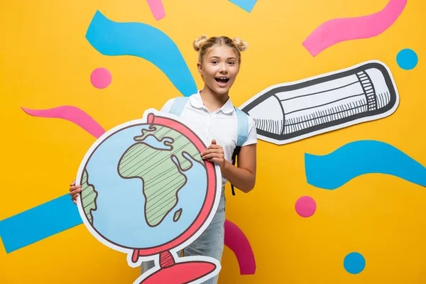 Excited schoolgirl holding globe maquette and looking at camera on yellow background with paper pencil and colorful elements — Stock Photo