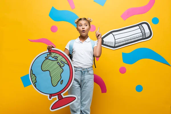 Colegiala sorprendida mostrando gesto idea mientras sostiene maqueta globo cerca de lápiz de papel y elementos de colores en amarillo - foto de stock