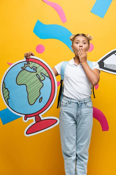 Шокована школярка покриває рот рукою, тримаючи макет глобуса біля паперового олівця та декоративних елементів на жовтому — стокове фото